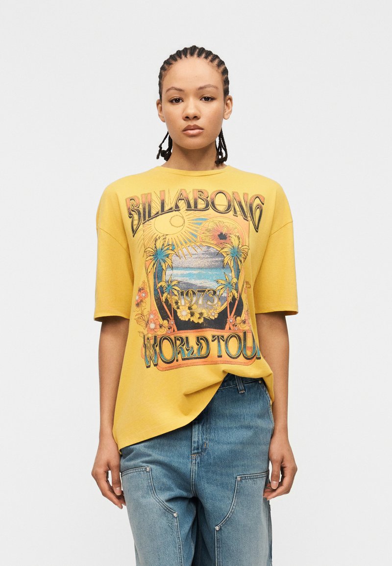 Γυναίκα με πλεξούδες που φοράει κίτρινο φαρδύ μπλουζάκι με πολύχρωμο σχέδιο «Billabong 1973 World Tour» και άνετο μπλε τζιν.