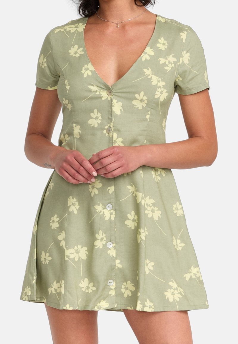 Femme vêtue d'une robe fleurie vert clair à manches courtes, col en V profond et boutons sur le devant, debout les mains jointes.