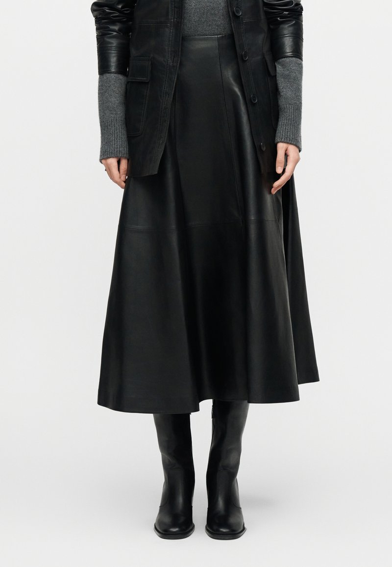 Γυναίκα φοράει μαύρη midi δερμάτινη φούστα, μαύρες μπότες μέχρι το γόνατο, γκρι πουλόβερ με μακριά μανίκια και μαύρο δερμάτινο μπουφάν, στέκεται μπροστά σε λευκό φόντο.