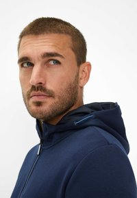 Felpa con zip di colore blu navy con finitura texturizzata, caratterizzata da un cappuccio con cordoncino e accenti tonali discreti. Tessuto morbido con una vestibilità aderente.