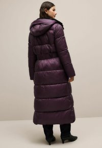 Pikk lill puffer-kate kapuutsi ja säästetud talje ning tikandiga osadega. Kangas on läikiv ja sile.