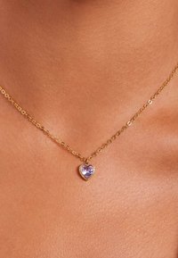 Gouden ketting met een hartvormige hanger met een paarse steen, omlijst door een parelrandontwerp, rustend op een lichte huidskleur.