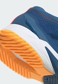 Modrá sportovní podrážka s výraznými oranžovými a bílými vzory pro záběr, texturovaná guma a elegantní, moderní design.