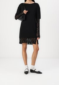 Zwarte jurk met kanten mouwen, kanten afwerking aan de zoom, gecombineerd met zwarte ballerina's met zilveren oogjes. Inclusief een kleine zwarte handtas.