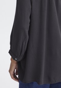 Black long-sleeve blouse featuring textured fabric, rolled cuffs with button detail, and a relaxed fit that extends below the waist.