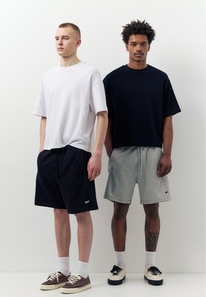 Dwaj mężczyźni stoją obok siebie, ubrani w casualowe szorty i obszerne t-shirty, jeden w biało-czarny, drugi w czarno-szary.