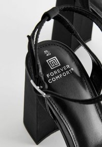 Black high-heeled sandals with a square heel, thin straps, and a cushioned insole. Branding visible on the insole with size details.