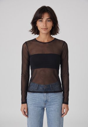 Mujer con cabello corto y oscuro, que lleva una blusa negra transparente de manga larga sobre un bandeau negro y vaqueros azul claro, de pie frente a un fondo liso.