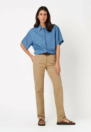 Woman standing with hand on hip wearing short-sleeve blue denim shirt, beige pants with belt, and brown sandals on white background.