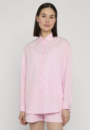 Mujer con cabello oscuro liso, vistiendo una camisa de manga larga con botones de rayas verticales en rosa claro y blanco, y pantalones cortos a juego con fondo liso.