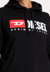 Personne portant un sweat à capuche noir avec un logo Diesel Denim Division rouge et blanc et une poche avant, montrée des épaules à la taille.