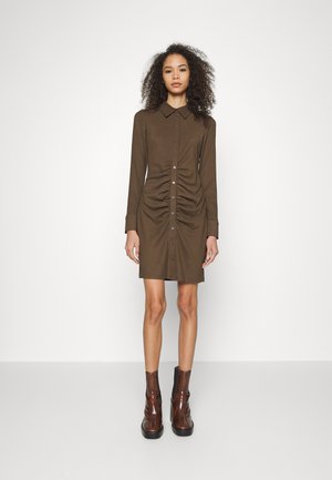 Vestido castanho com botões e mangas longas, apresentando detalhes franzidos nas laterais. Combinado com botas castanhas brilhantes até ao joelho.