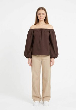 Kvinde stående iført en brun off-shoulder bluse med lange ærmer, beige vidde bukser og hvide sneakers på en hvid baggrund.