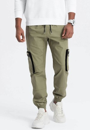 Homme portant un pantalon jogger cargo olive clair avec des poches zippées noires, des baskets blanches, un sweat-shirt blanc et une montre-bracelet blanche, debout.