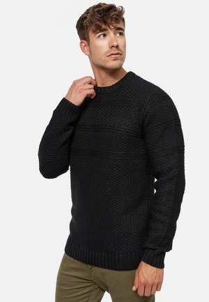 Suéter negro de punto con un patrón texturizado, puños y dobladillo acanalados. Escote redondo con sutiles rayas horizontales. Combinado con pantalones verdes.