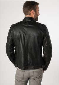 Schwarze Lederjacke mit strukturiertem Design, strukturierter Oberfläche, aufgerauten Schulterakzenten und gepolsterten Ellenbogenbereichen. Graue Jeans runden den Look ab.