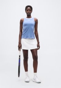 Γυναίκα αθλήτρια με μπλε αμάνικο τοπ και λευκή φούστα, κρατώντας ρακέτα τένις, στέκεται μπροστά σε απλό λευκό φόντο.