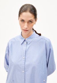 Chemise à boutons rayée bleu clair et blanche avec col, présentant une coupe loose et une texture de tissu douce. Détails des boutons et des poignets visibles.