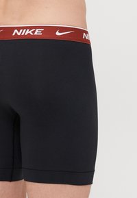 Sorte boxer briefs med en mørkerød elastisk talje, der har et hvidt "Nike" logo. Glat stof med en tætsiddende design og flade sømme.