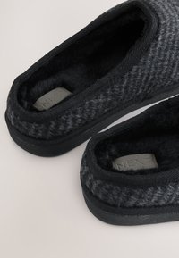 Chausson noir avec une tige texturée, une doublure en fourrure, et une semelle en caoutchouc noire. Présente des bords cousus et une étiquette grise avec « NEXT » à l'intérieur.