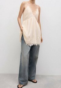 Camisola beige con ribete de encaje y tirantes finos, con un ajuste suelto. Combinada con vaqueros de pierna ancha de color azul claro y sandalias negras.