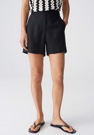 Vrouw die zwarte, op maat gemaakte shorts met hoge taille draagt, een zwart-wit patroonloze mouwloze top en zwarte teenslippers, staand met één hand in de zak.