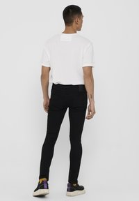 Camiseta blanca de manga corta, jeans negros ajustados, zapatillas negras con acentos morados y amarillos, vistas desde atrás contra un fondo liso.