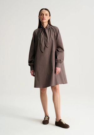 Kobieta stoi ubrana w brązową, kraciastą sukienkę z długim rękawem i kokardką na szyi oraz brązowe trampki, na jednolitym jasnym tle.