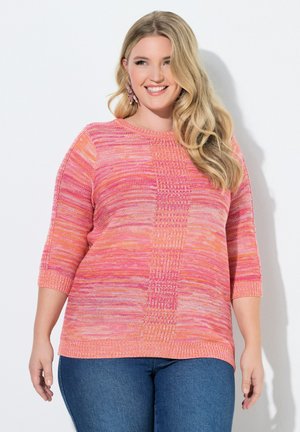 Smilende kvinde med langt blondt hår iført en pink og orange strikket sweater og blå jeans, stående mod en hvid baggrund.