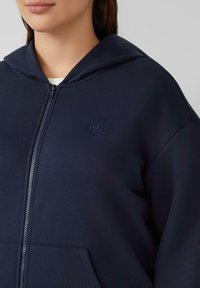 Marineblauwe hoodie met rits, uitgevoerd in een textuurstof, met voorzakken en een subtiele geborduurde logo op de borst.