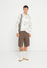 Felpa grigia con motivo floreale e grande logo, pantaloni cargo marroni con tasche laterali, sneakers beige e calzini bianchi, su uno sfondo neutro.