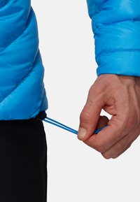 Veste isolante bleue avec un design matelassé. Main ajustant un cordon de serrage à la taille. Tissu texturé, accent noir près du cordon.