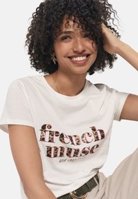 Weißes Baumwoll-T-Shirt mit dem Text "French Muse" in Leopardenmuster und pinken Akzenten. Das Model posiert mit lockigem Haar, trägt goldene Schmuckstücke und beige Hosen.