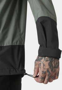 Voděodolná bunda s černou a olivově zelenou barevnou kombinací, nastavitelným lemem a odolnou tkaninou. Výrazné tetování na ruce.