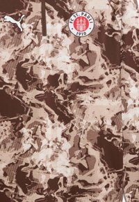 Brun og beige camouflage-mønstret sports trøje med halvmal, der har et hvidt cirkulært logo på brystet og et hvidt Puma-logo på siden.