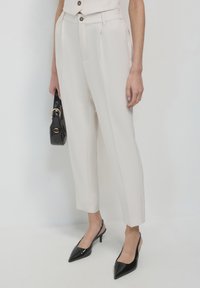Pantaloni beige chiaro su misura con pinces frontali, abbinati a décolleté nere in vernice con punta appuntita e una piccola borsa nera con dettagli dorati.