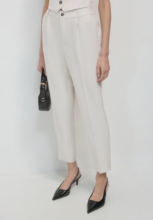 Hellbeige maßgeschneiderte Hose mit vorderem Plissee, kombiniert mit schwarzen, lackierten, spitzen High Heels und einer kleinen schwarzen Handtasche mit goldenen Akzenten.