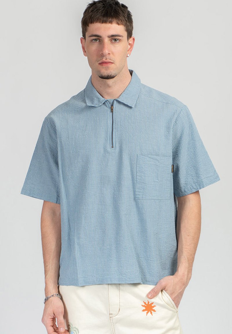 Νέος άνδρας που φοράει ένα ανοιχτό μπλε κοντομάνικο υφασμάτινο πόλο με εμπρός τσέπη και κρεμ παντελόνι με πορτοκαλί σχέδιο ήλιου.