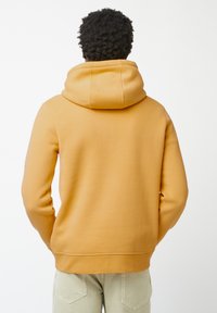 Felpa con cappuccio in giallo senape, realizzata in tessuto morbido. Dotata di tasca a marsupio e polsini a coste, presenta una vestibilità comoda e una consistenza liscia.