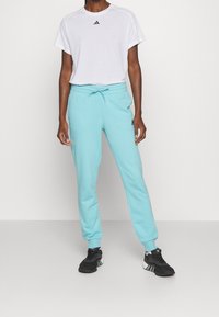 Person klädd i vit T-shirt med kort ärm och ljusblå Adidas sportbyxor, som står på en slät grå golvyta med svarta Adidas sneakers.