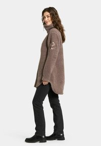 Fleece pullover i taupe med høj krave, sideslids og en struktureret overflade. Båret med sorte jeans og ankelstøvler.