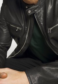 Černá kožená bunda s hladkou texturou, s stříbrn�ými zipy a cvočky. Nošena přes tmavě zelenou košili.