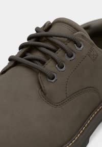 Braunes Wildleder-Schuhwerk mit sechs Metallösen, dunkelbraunen Schnürsenkeln und dezenten Steppdetails. Runde Zehenpartie und strukturierte Oberfläche.