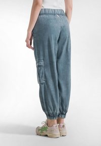 Jasnoniebieskie bawełniane spodnie bojówki z elastycznymi mankietami, wyposażone w boczną kieszeń, gładką fakturę i luźny krój. Noszone z beżowymi sneakersami.