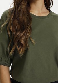 Lang, bruin, golvend haar over een groene shirt met korte mouwen, elastische manchetten en een ronde hals op de bovenkant van een persoon.