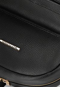 Zaino in pelle nera di texture con forma rotonda, caratterizzato da accessori in oro e un marchio ben visibile. Dettaglio della chiusura con zip in evidenza.
