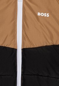 Giacca puffer marrone e nera con zip bianca, dotata di un prominente logo "BOSS" nella parte superiore. Tessuto liscio e lucido.