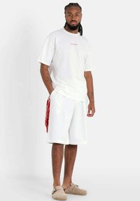 Witte T-shirt met korte mouwen en rode tekst, gecombineerd met losse witte shorts en beige instapschoenen, staand tegen een effen achtergrond.