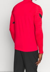 Camisa deportiva de manga larga en un rojo brillante con mangas texturizadas, que presenta detalles en negro cerca de los hombros y un diseño ajustado.