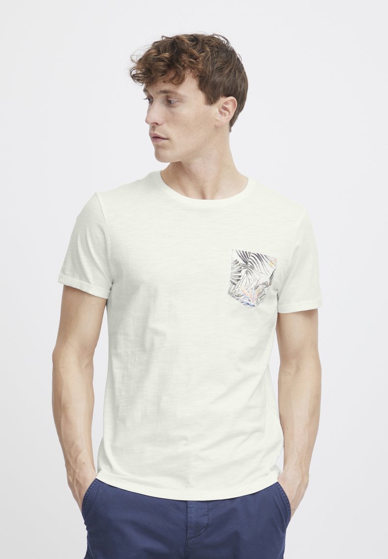 Camiseta de color beige claro con mangas cortas, que presenta un bolsillo en el pecho con un patrón de hojas tropicales. Textura suave y corte relajado.